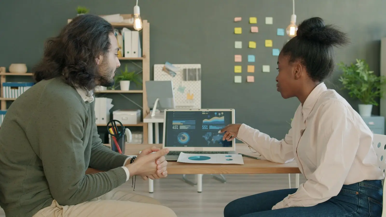 Two colleagues at a desk discussing charts on a laptop screen